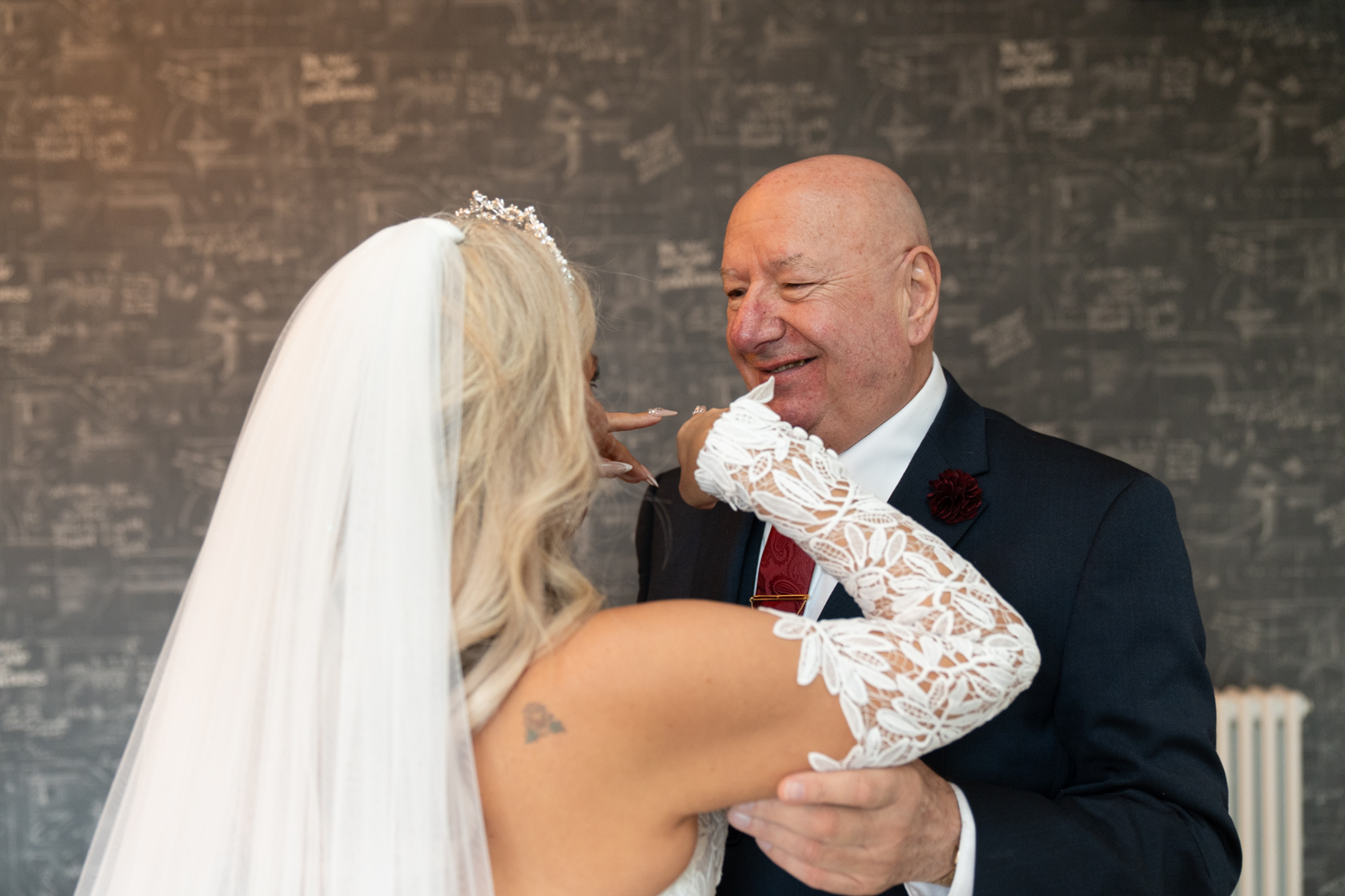 The bride in her dress and veil reaches for her father's face — he's beaming, barely holding it together, the first time he's seen her